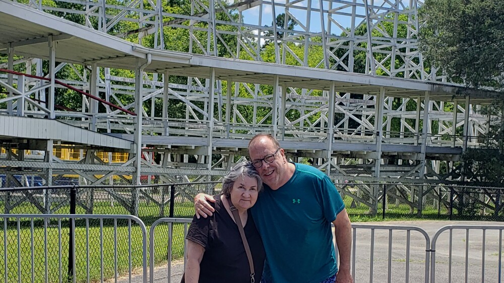 A smiling couple poses warmly in front of a large wooden roller coaster under a bright blue sky. Their casual attire suggests a day of leisure and fun.