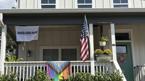 A white house porch with a 'Highland Park Porchfest' banner and an American flag. A vibrant Pride flag hangs on the railing, surrounded by plants.