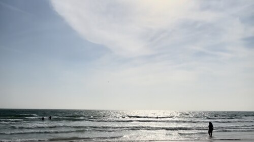 The sun shines at the beach. One person is visible in the shallows of the ocean, and waves are lapping onto the sand.