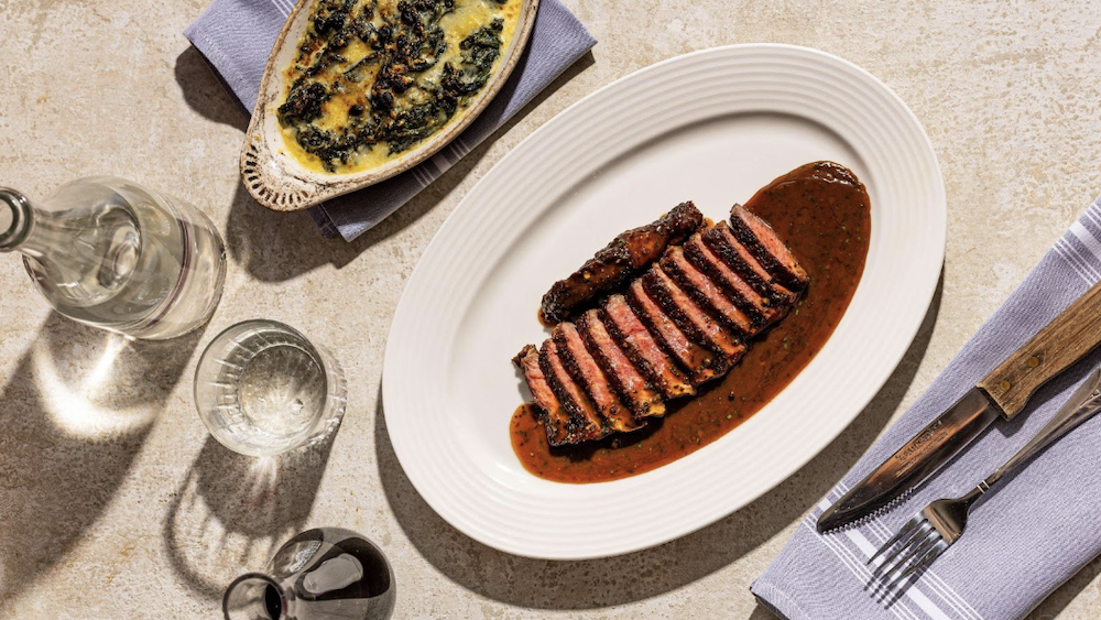 A plate containing charbroiled steak au poivre, next to silverware, glassware, and a dish of creamed spinach.