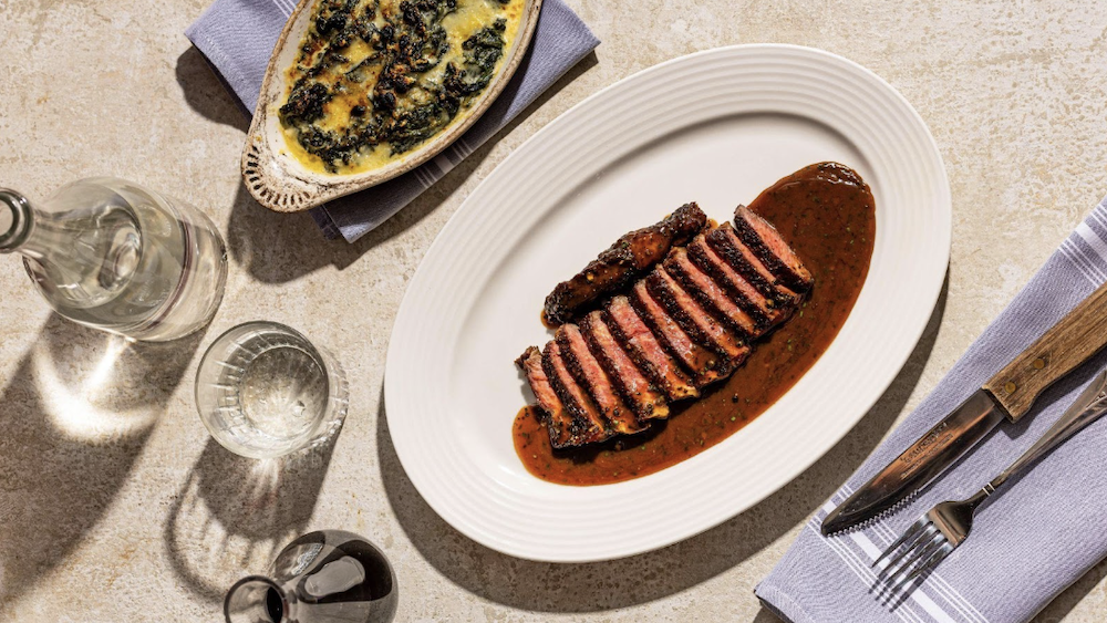 A plate containing charbroiled steak au poivre, next to silverware, glassware, and a dish of creamed spinach. 