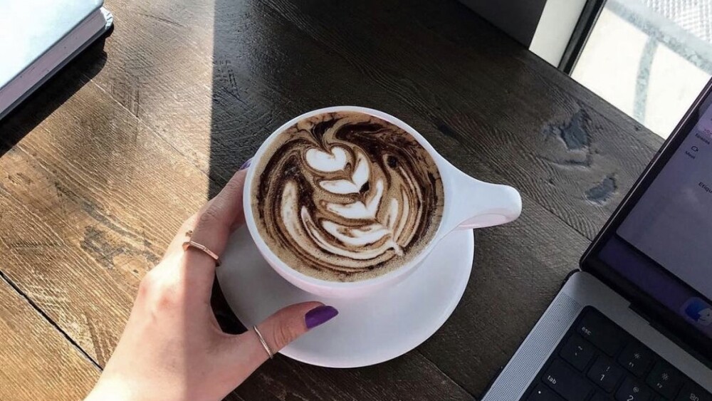A hand with purple painted nails and two small-band gold rings lovingly caresses a cup with ornate latte art, dangerously close to a laptop.
