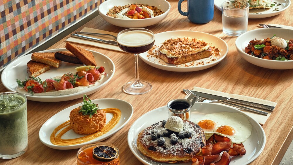 A vibrant brunch spread displayed on a wooden table, featuring dishes like blueberry pancakes with bacon and eggs, smoked salmon with toast, granola with fresh fruit, a sweet potato hash, and a frosted slice of cake. Drinks include an espresso martini, matcha latte, black coffee, and a cocktail garnished with a dried citrus slice. A colorful checkered booth lines the back of the table.