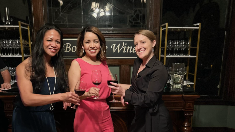 Three people standing with wine glasses. The two people dressed in black are on either side of a person in a pink jumpsuit in the middle. A wine rack and mirror is behind them.