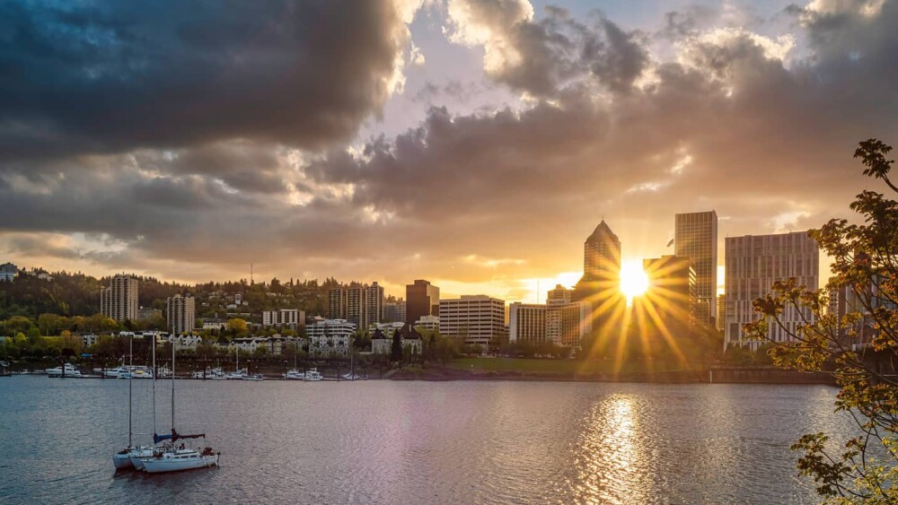 The sun shines between two tall buildings in downtown portland, with the river in the foreground, under a stormy sky