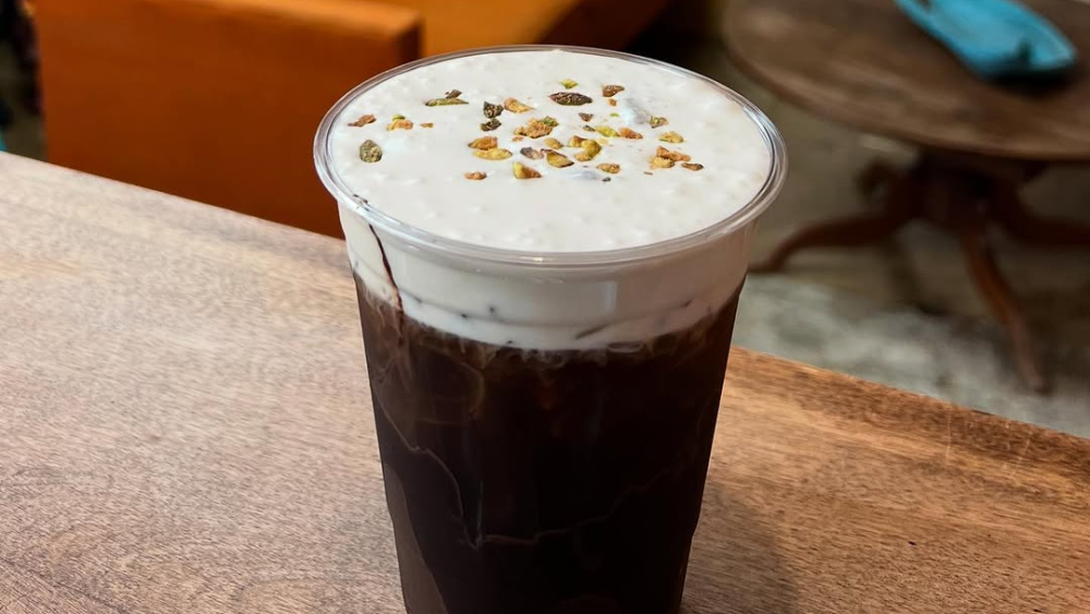 Dubai chocolate Cold Brew from Pour Bear Coffee topped with cold foam and crushed pistachios, set on a wooden counter near a cozy couch.
