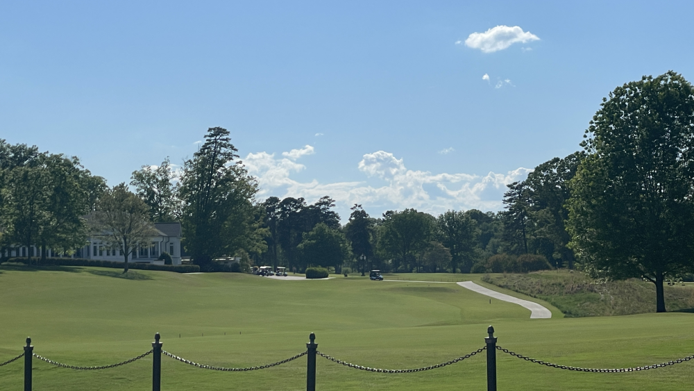 Green hills with trees. A white clubhouse is in the background and golf cars are driving around. A black fence is in the foreground.