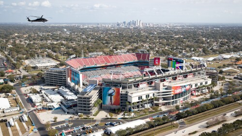 A helicopter flies over Raymond James Stadium before Super Bowl LV. Tampa's skyline is visible in the background.