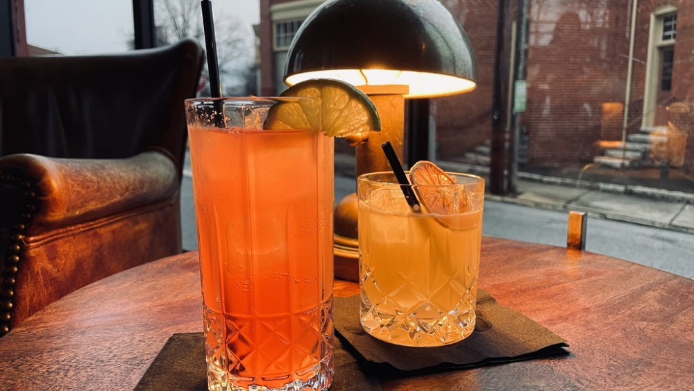 Tall pink drink with lime slice and small orange cocktail with burnt orange slice. Both are sitting on black napkins on a wooden table and windows are behind them.