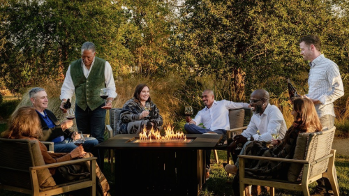 A group of people enjoy glasses of wine around a fire pit at Argyle Winery. The scene is set by verdant trees in the background. Golden light suggests a time just before sunset.