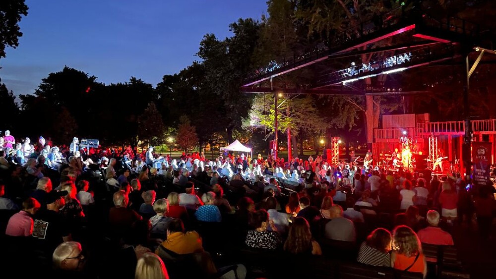A concert stage is lit with red light, while a band performs before a crowd of a few hundred people.