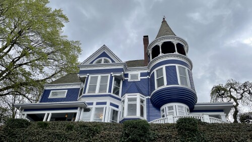 A Queen Anne style mansion painted dark blue with white trim, a conical current topped by a porch, and bay windows.