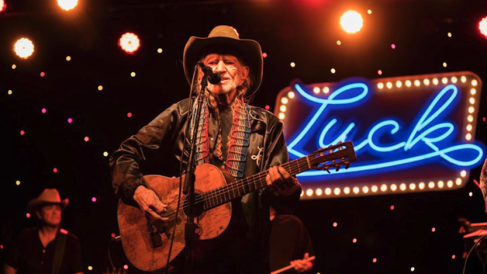 Willie Nelson playing guitar in front of a neon sign reading "Luck."