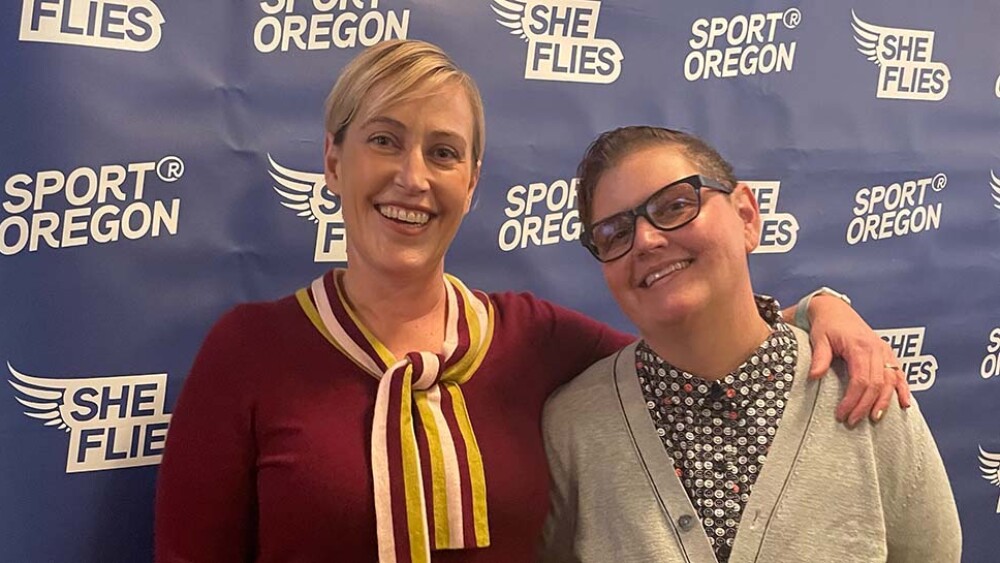 Two people stand side by side for a photo in front of a photo backdrop with the words "Sport Oregon" and "She Flies"