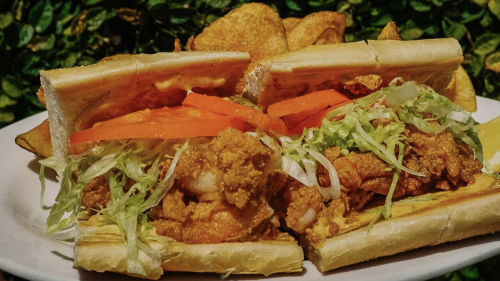 A fried shrimp po boy at Willa's in Tampa. The sandwich is on a baguette, with battered shrimp accompanied by sauce, lettuce, and tomato, with potato chips also on the plate in front of a green leafy backdrop.