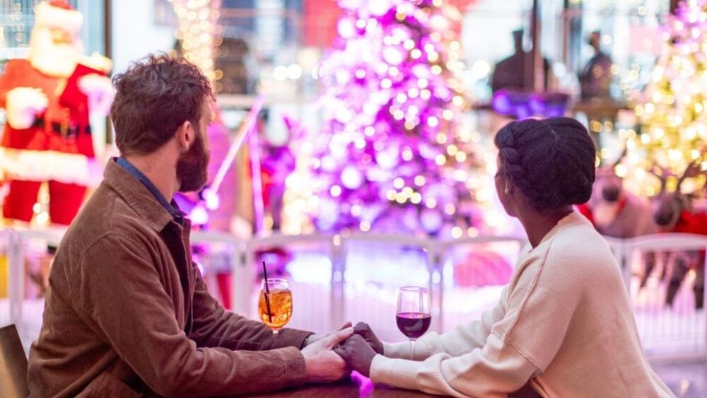 A couple sitting at a table with drinks, holding hands and looking at a Christmas light display in the background.