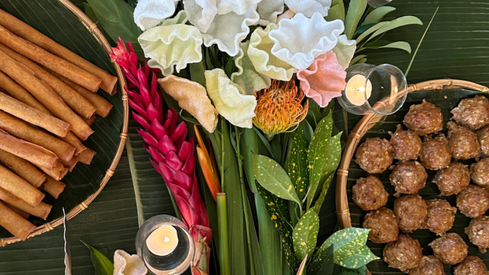 An overhead spread features golden lumpia, savory meatballs, colorful fried shrimp chips, and tropical flowers on banana leaves. Two small candles glow warmly, enhancing the vibrant, festive, and inviting presentation.