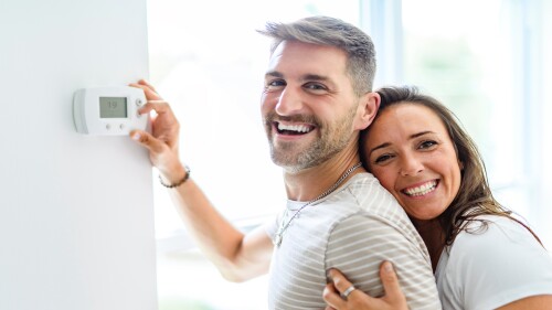 A smiling couple embracing while the person in front holds the thermostat.