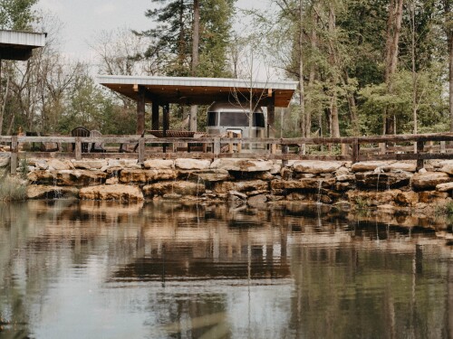 An Airstream trailer sits beneath a roof near a stone-lined pond.