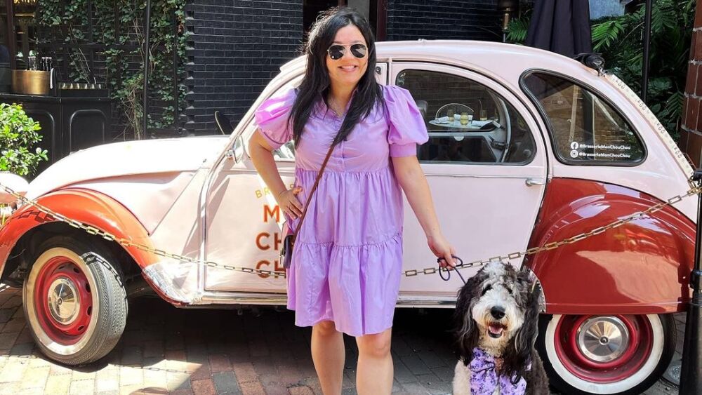 A person wearing a purple dress and a dog on a leash wearing a matching purple bow stand in front of a displayed vehicle.
