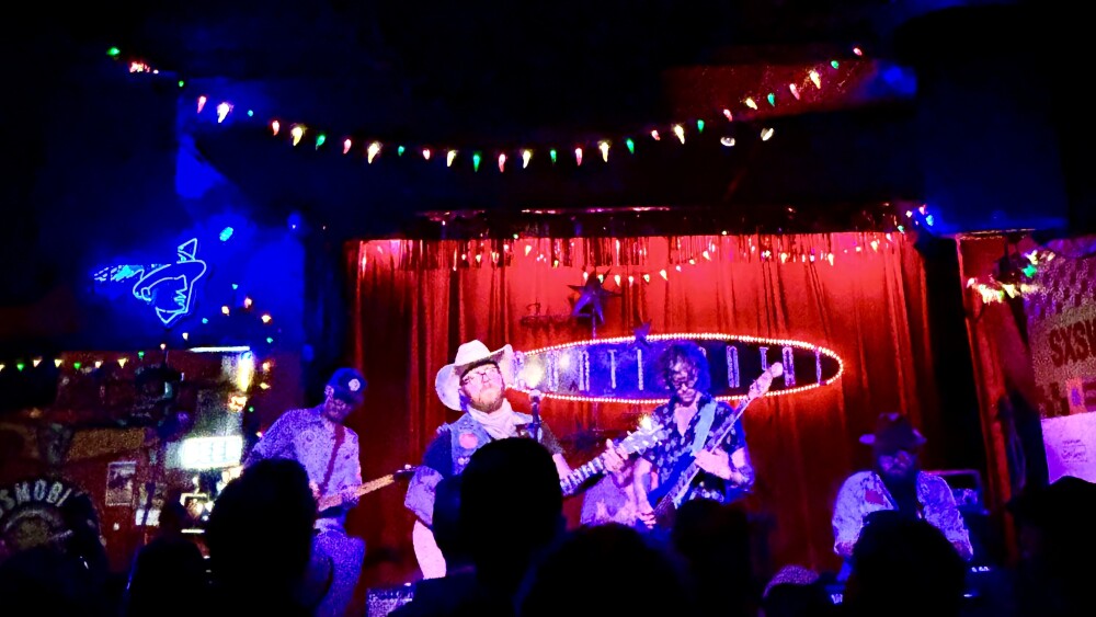 performer on a stage in a cowboy hat, the stage has red draping and twinkly lights