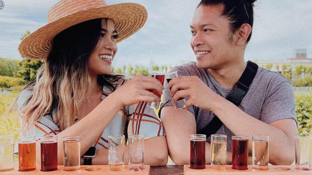 Two people cheers their tasting glasses of cider at an outdoor picnic table.