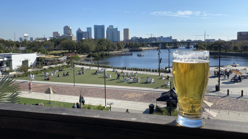 A mostly full glass of beer at Armature Works sits on a second-floor ledge. People are chilling by the river front below, and the blue sky has a handful of wispy clouds above the distant Tampa city skyline.