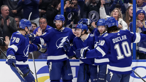 A group of Tampa Bay Lightning players celebrates a goal by the glass. They are dressed in their blue uniforms and are hugging each other while the crowd celebrates behind.