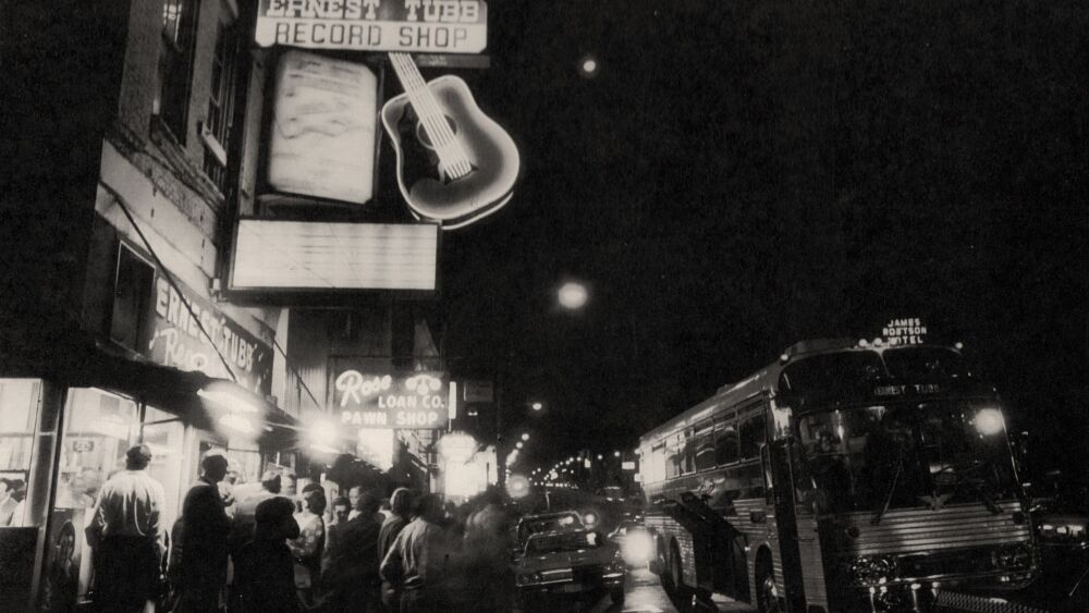 A black and white image of the exterior of the former Ernest Tubb Record Shop at night. A crowd of people stand under its marquee sign as a bus drives by down Broadway.
