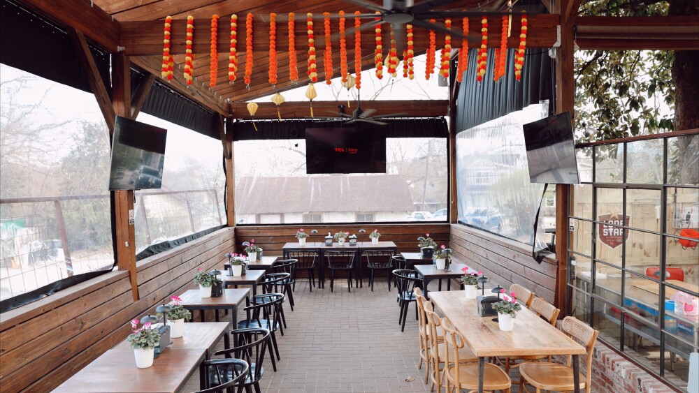 Wooden bench seating lines the left, back, and partial right wall with nine tables. A longer table sits on the far right lined on either side with six chairs total. Three TVs hang on the wall and the space is fully enclosed.