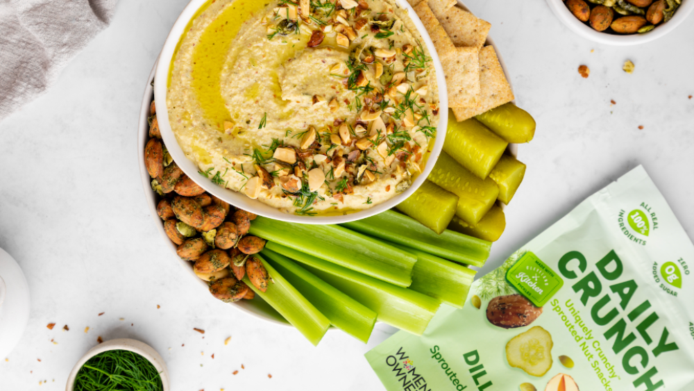A platter with dip topped with Daily Crunch nuts, pickles, celery, chips, and more Daily Crunch snacks sits on a table next to a bag of the product.