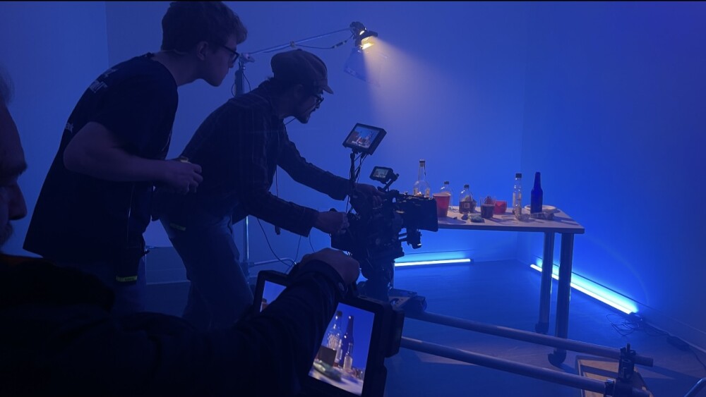 A cameraman with a tight shot of bottles surrounded by other production people in a room with blue lights.