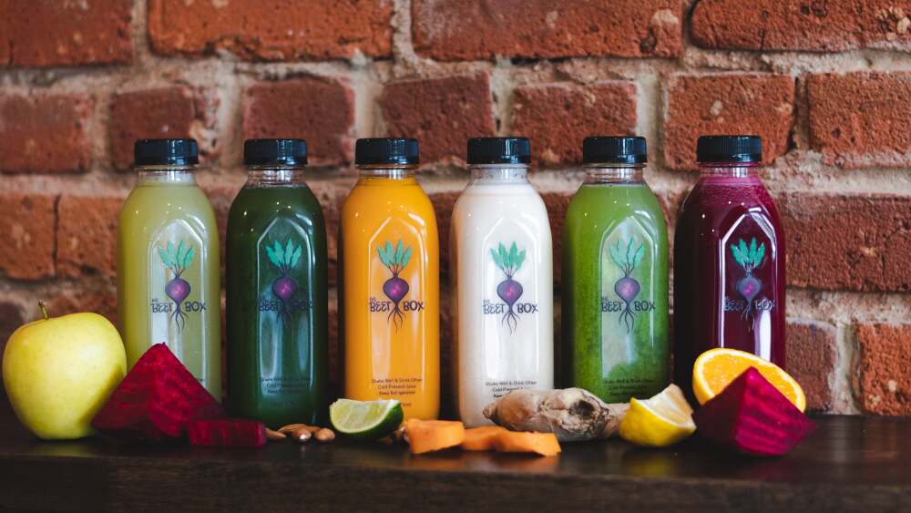 Six juices sit on a table in front of a brick wall.
