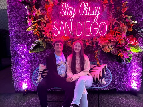 A couple poses under a neon sign that reads "Stay Classy San Diego."