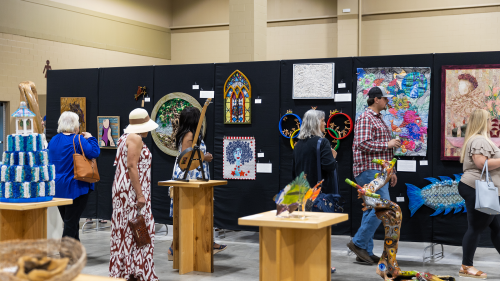 Visitors browse a vibrant indoor art exhibit showcasing mixed media, sculptures, and colorful wall hangings. People of various ages engage with the artwork, some admiring stained glass, fiber art, or abstract pieces. Sculptures of animals and intricate textures create a lively, creative atmosphere within a spacious, well-lit venue.