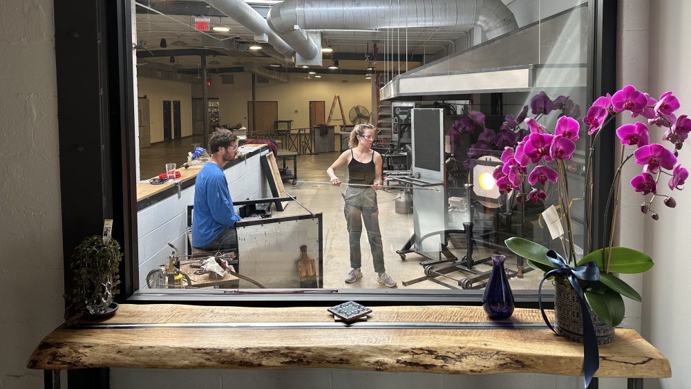 A view through a window to the workshop where two artists are blowing glass.