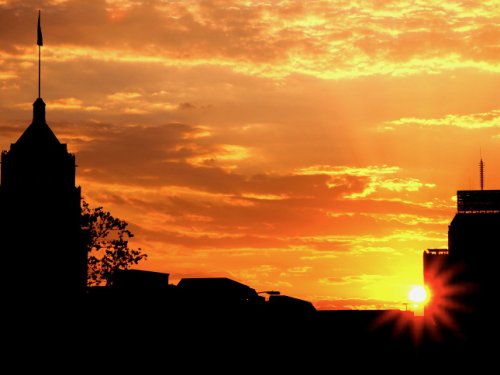 The sun setting behind the San Antonio skyline. An orange sky with wispy clouds is behind several silhouetted buildings.