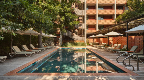 A rectangular pool is surrounded by wooden decking and lounge chairs shaded by white umbrellas, nestled beneath mature trees and bordered by red brick walls. In the background, a mid-century modern style building with a red and gray patterned facade rises above the scene, casting shadows across the quiet, shaded courtyard.