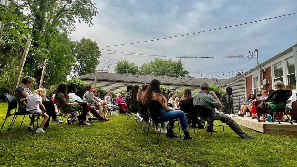 A group of people sitting in chairs on a lawn listening to a group of people speak.