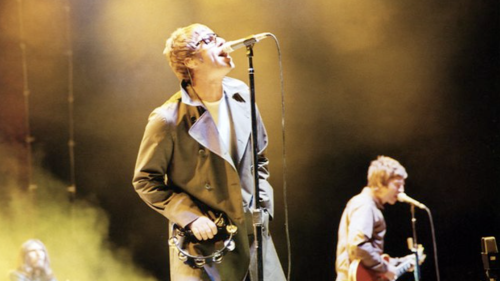 The Gallagher brothers perform with Oasis. There is yellow smoke and lighting around while they sing into microphones and play instruments.