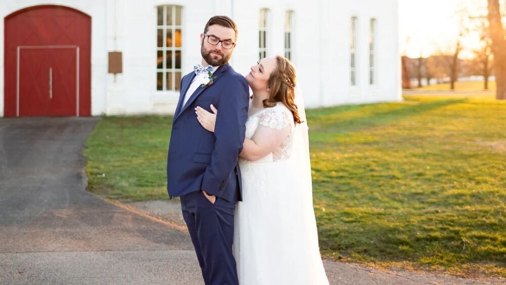 A couple in front of a white round barn with a red door.