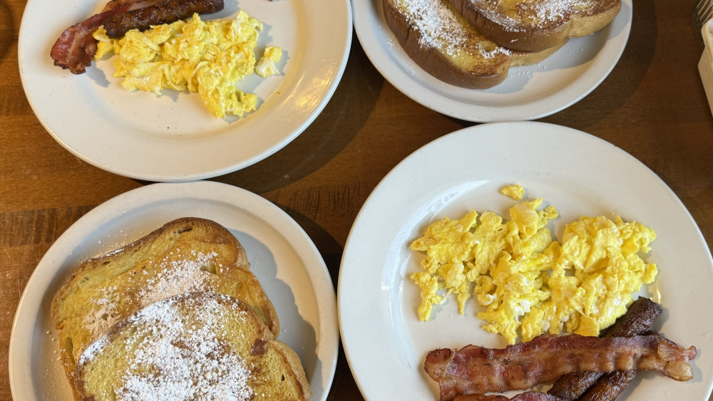 Four white plates sit on a brown wooden table. Two contain two slices of French toast while the other two contain scrambled eggs, bacon, and sausage links.