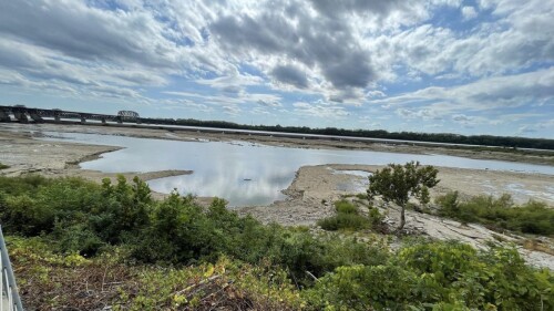 The river level is low so the riverbed is exposed with green foliage, rocks and sticks. In the distance, you can see a defunct railroad trestle.