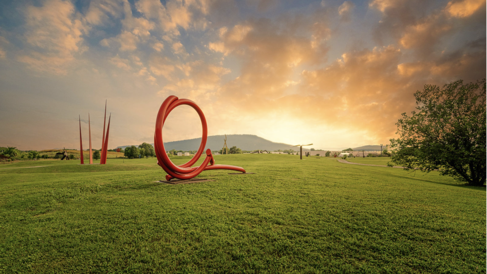 Sunset over a grassy field with vibrant red abstract sculptures. The sky features dramatic clouds, adding a serene yet dynamic atmosphere.
