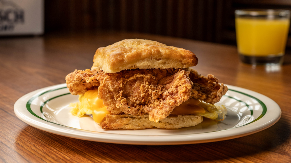 A plate with one biscuit sandwich made with fried chicken, egg, and melted cheese and a glass of orange juice out of focus in the background.