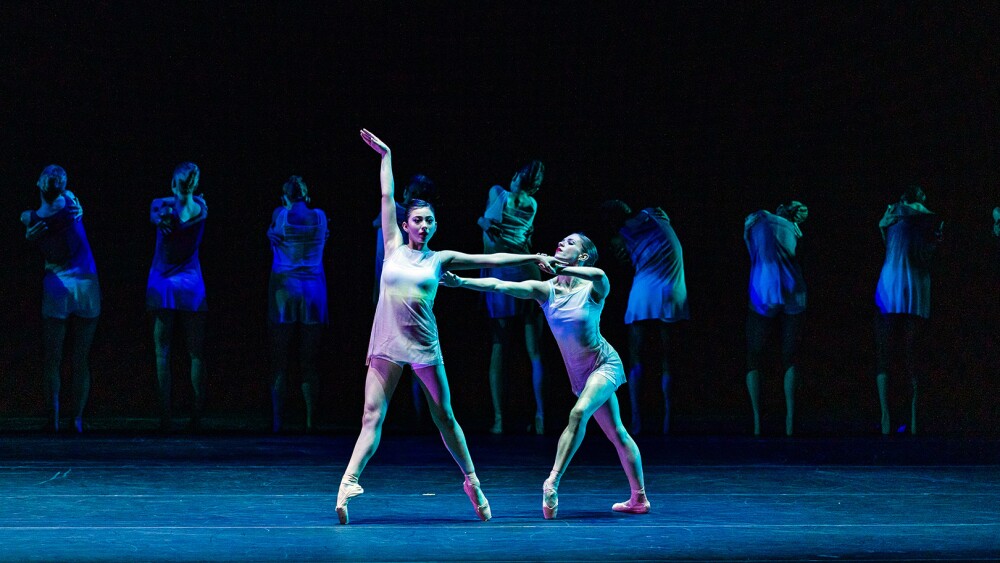 Two dancers stand onstage in the spotlight with shadowy dancers behind them.