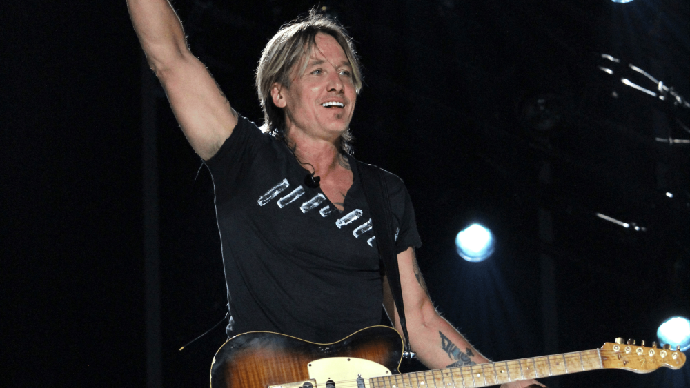 Keith Urban stands on stage at CMA Fest with one arm holding a mic in the air and the other on his guitar.