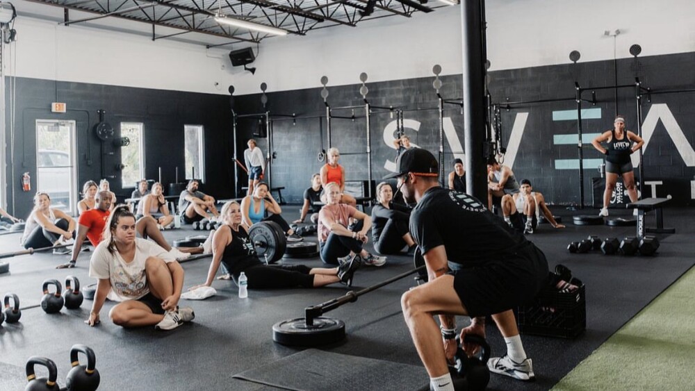 A class at Sweat St. Pete. There is a black mat with people training, sitting facing an instructor who is demonstrating a workout with kettlebells.