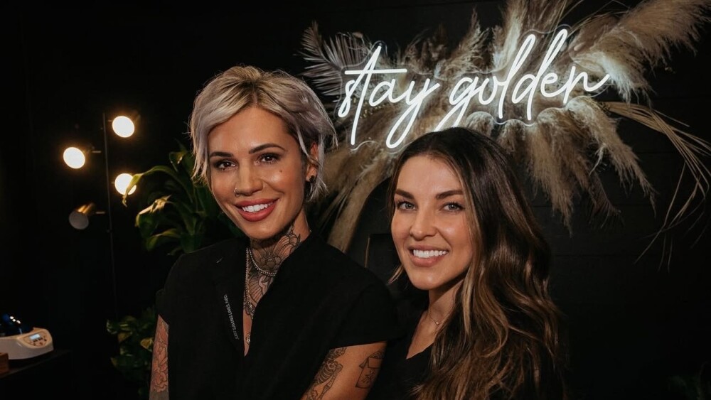 two women pose in front of a neon sign that says stay golden