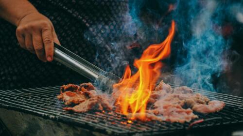meat cooking on a grill with flames coming up and a person's hand in the top left corner